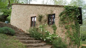 Un bâtiment en pierre avec deux fenêtres, un escalier menant à l’entrée et des fleurs blanches qui poussent autour.