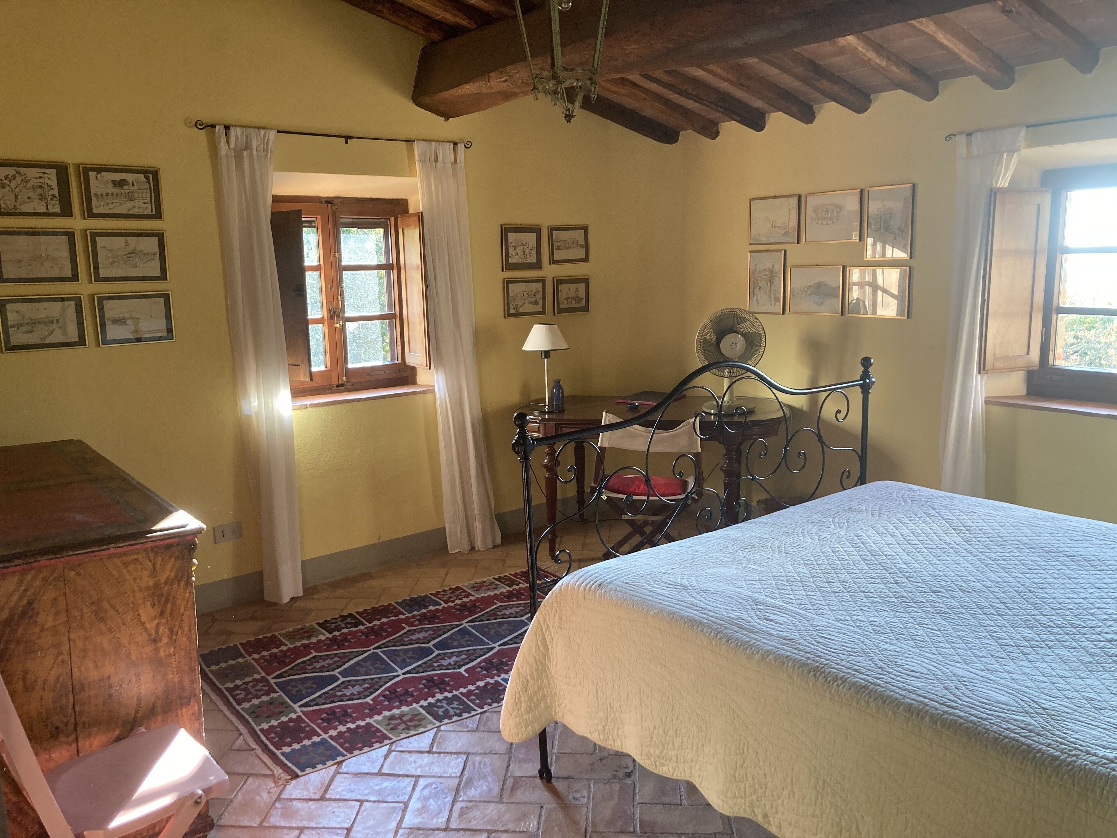 Une chambre à coucher comprenant un lit, une commode en bois, un bureau avec une chaise, une fenêtre avec des rideaux et plusieurs tableaux encadrés accrochés aux murs.