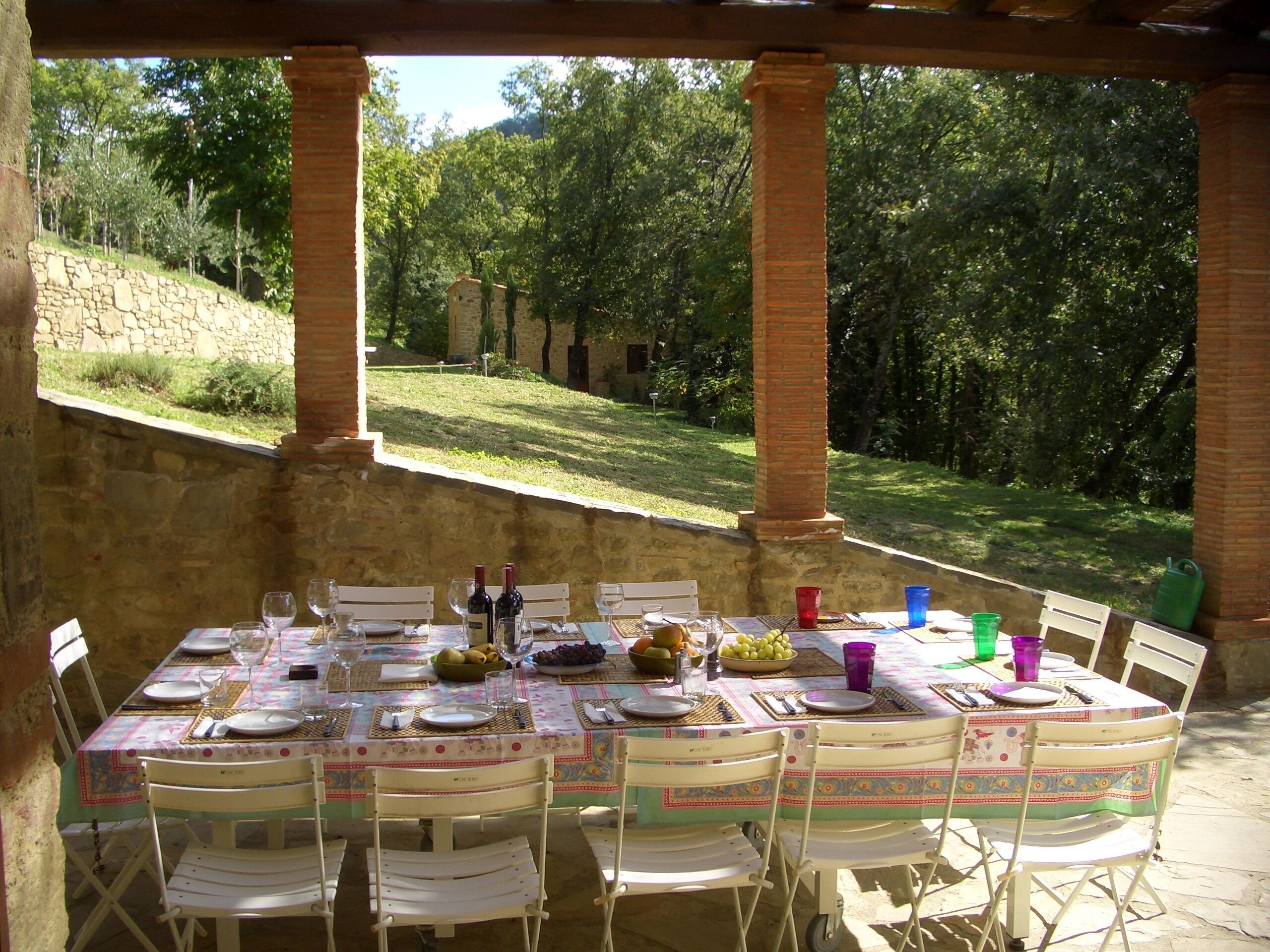 Un área de comedor al aire libre con una mesa dispuesta para seis personas, rodeada de sillas blancas, bajo una estructura de ladrillo con vistas a una colina cubierta de césped y árboles.