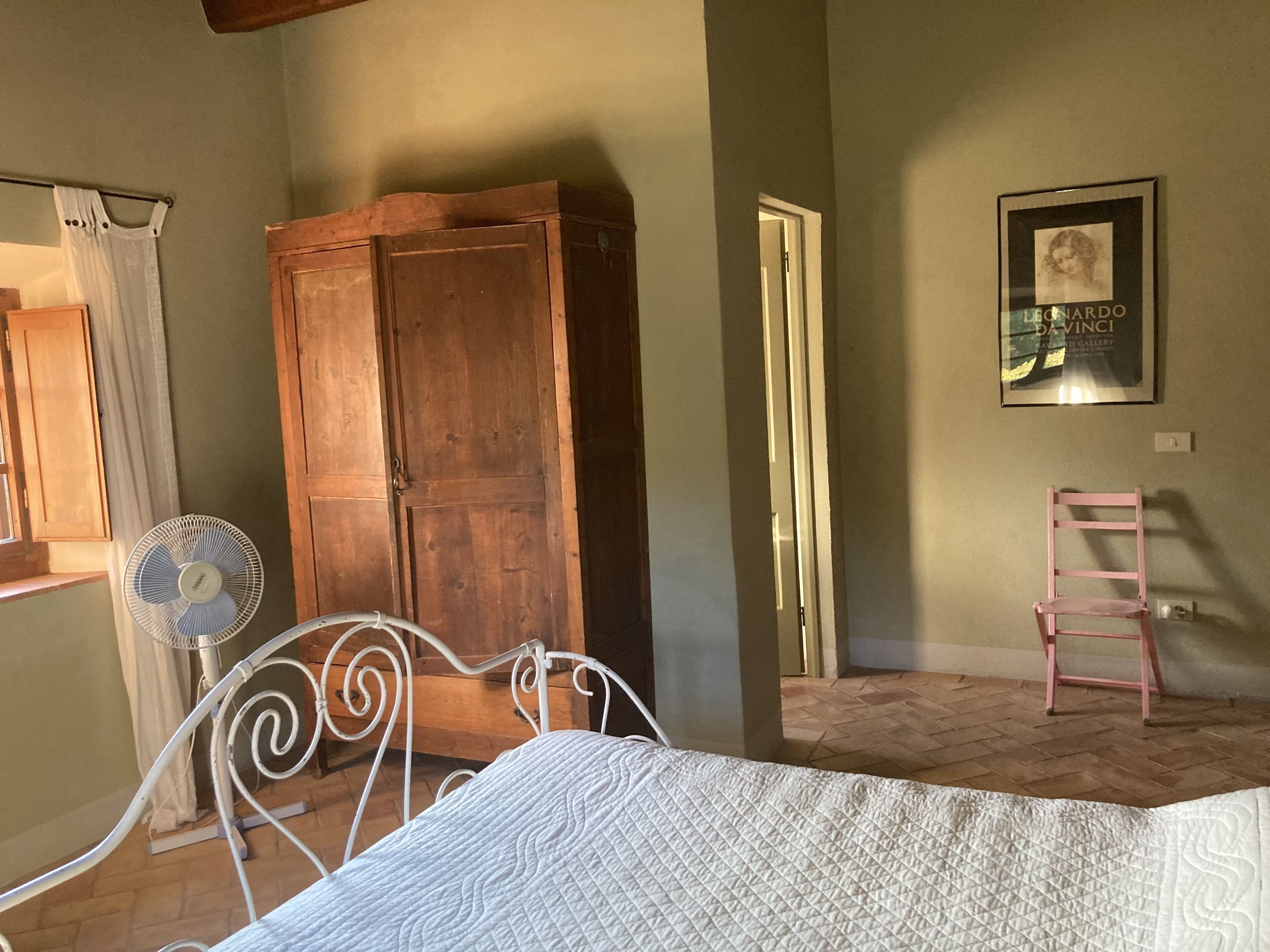A bedroom with a bed, a wooden wardrobe, a standing fan, a chair, and a framed picture on the wall.