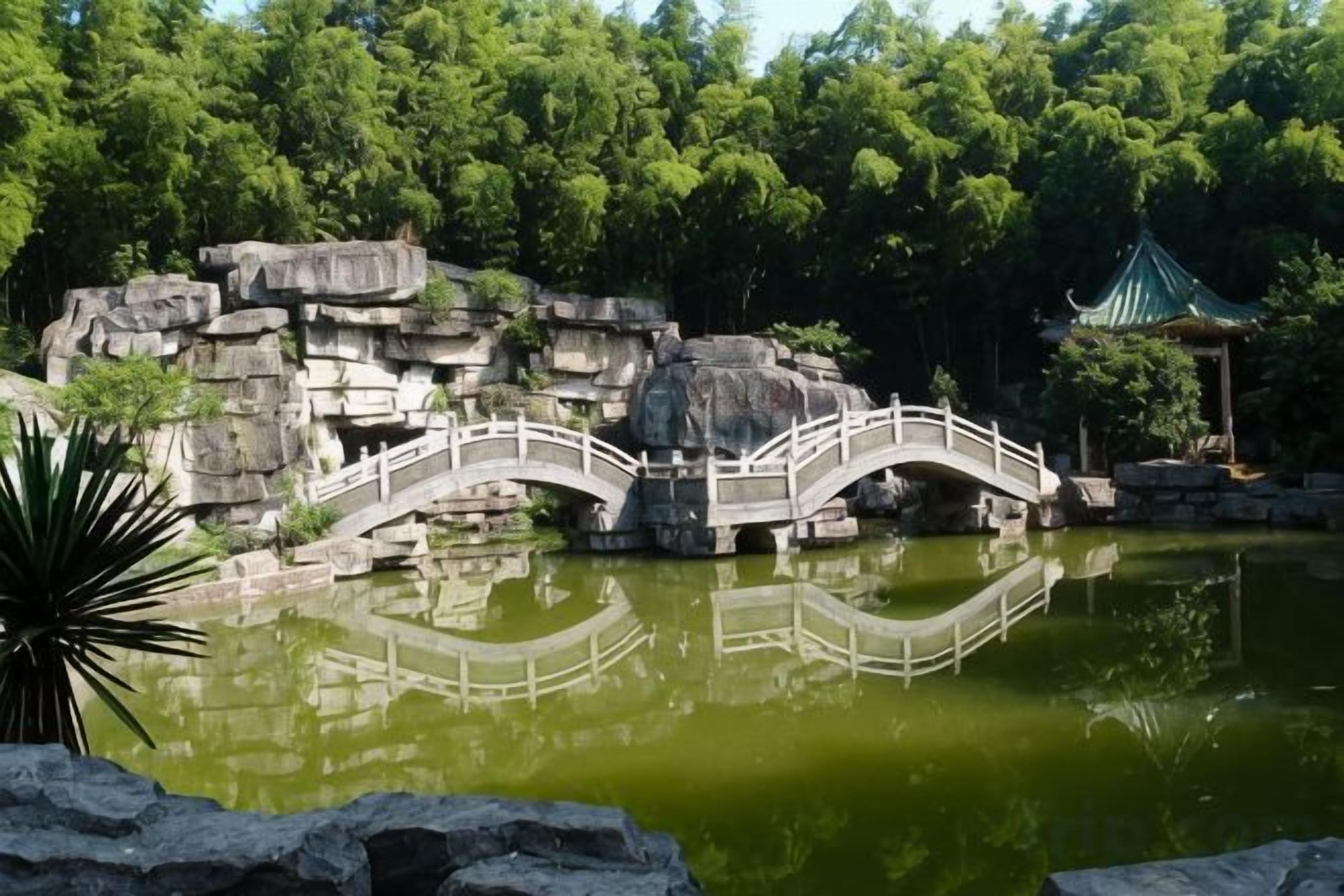 A bridge over a pond with a rock formation and a traditional pavilion in the background.