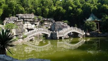 A bridge over a pond with a rock formation and a traditional pavilion in the background.