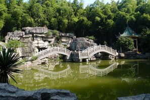 A bridge over a pond with a rock formation and a traditional pavilion in the background.