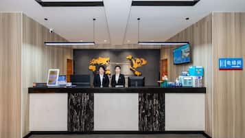 A hotel reception desk with a flat-screen TV, a digital clock, and a sign indicating a self-service kiosk.