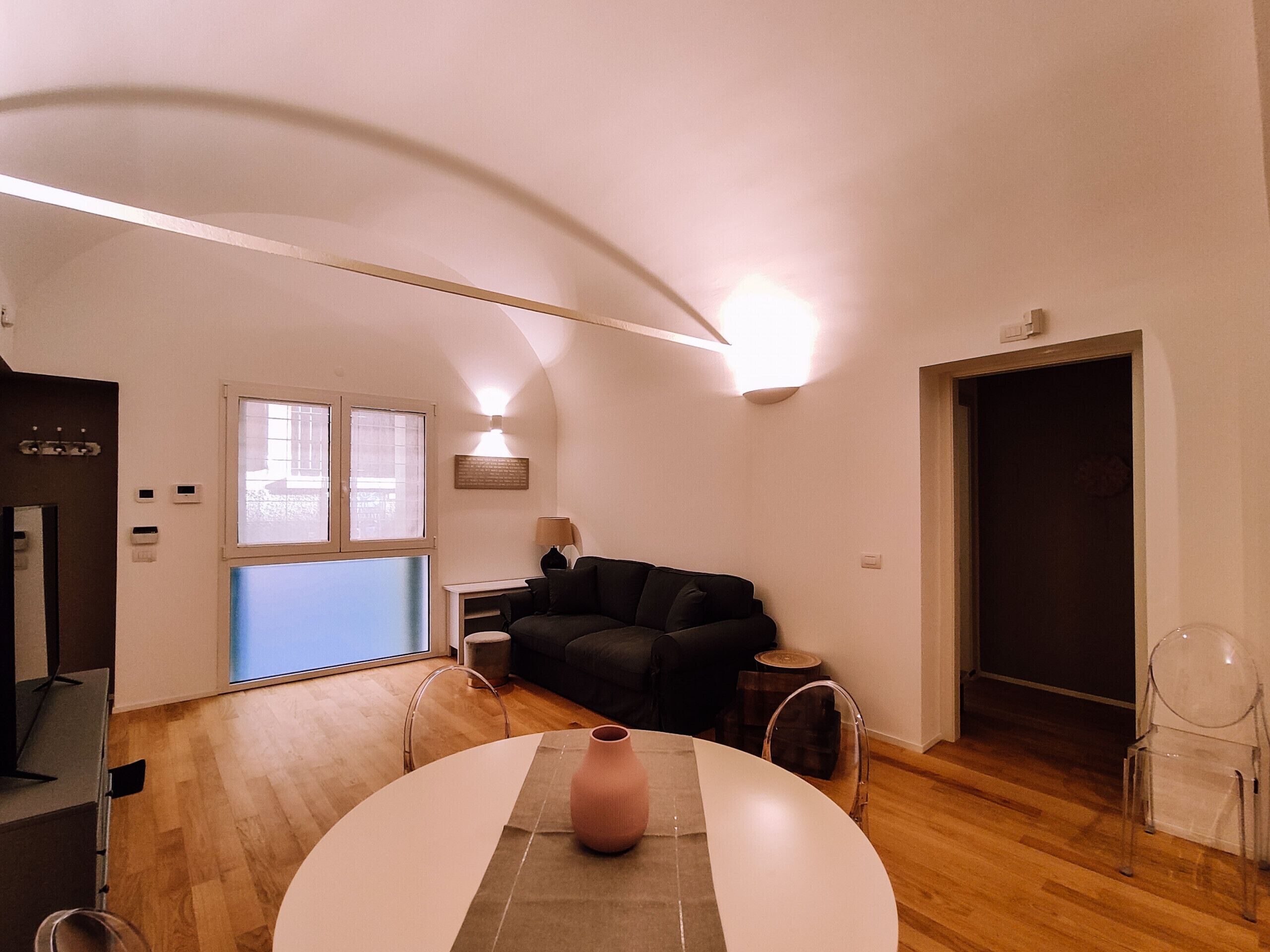 A modern living room with a curved ceiling, a round table with a vase, a black sofa, and a clear chair.