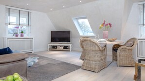 A modern loft bedroom with a sloped ceiling, a large skylight, a flat-screen TV, wicker furniture, and a dining area. - large modern beach house style apartment (Sylt)