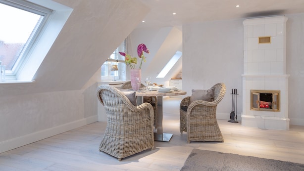 A modern loft bedroom with a sloped ceiling, a fireplace, and a dining area with wicker chairs and a table. - large modern beach house style apartment (Sylt)