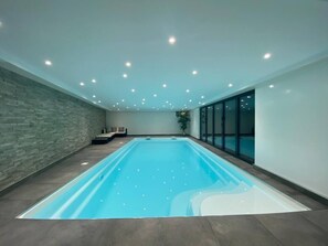 A large indoor swimming pool with a stone wall and a bench, surrounded by a glass enclosure.