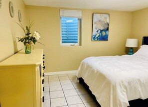 A bedroom with a bed, a dresser, a window with blinds, a painting, and a vase with flowers.