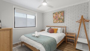 A bedroom with a bed, a wooden dresser, a window with blinds, a ceiling fan, a brick wall, and a painting. - Orion Retreat by Experience Jervis Bay (Vincentia)