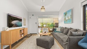 A modern living room with a flat-screen TV, a wooden cabinet, a grey sofa, a blue armchair, and a large window with a view of greenery. - Orion Retreat by Experience Jervis Bay (Vincentia)