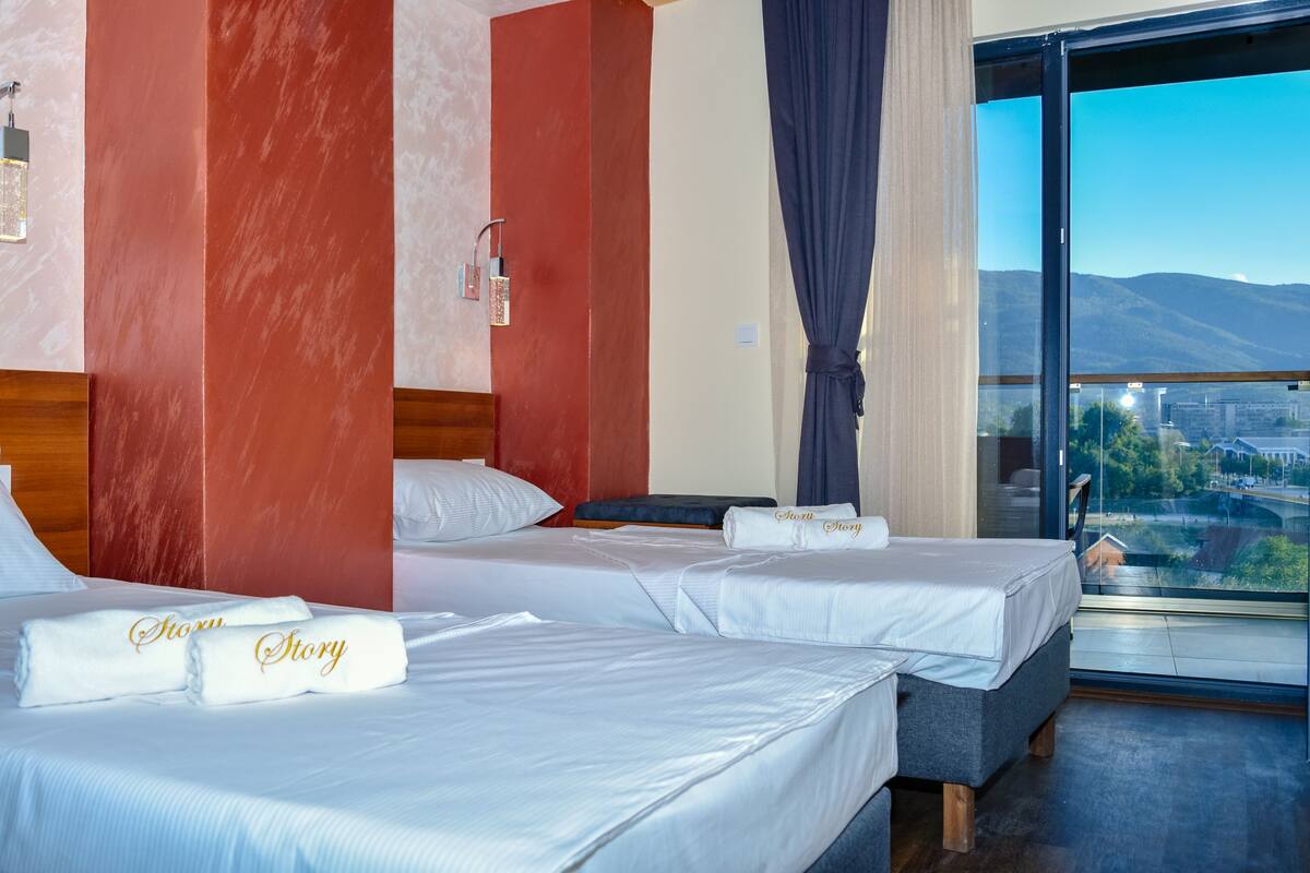 Habitació d'hotel amb dos llits, un gran finestral amb vista a la ciutat i una paret vermella decorativa.