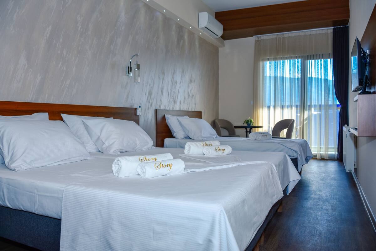 Habitació d'hotel amb dos llits, cada un amb roba de llit i tovalloles blancs, amb la paraula 'Story' escrita. Hi ha una taula de treball amb una cadira, una taula petita amb una planta i una finestra amb cortines.