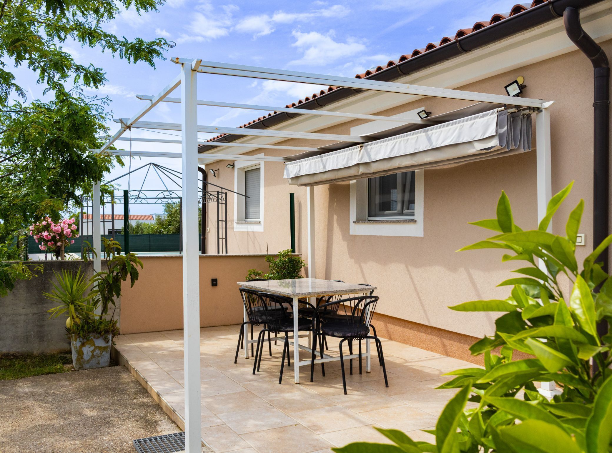 An outdoor patio with a table and chairs, a pergola, and a view of a building with a tiled roof.