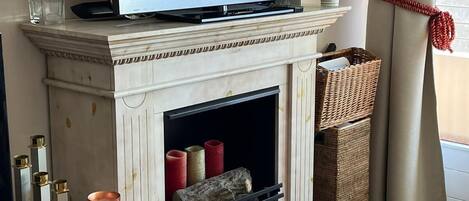 Un salon moderne avec un téléviseur à écran plat posé sur une cheminée, un foyer avec des bûches et une vue sur l’extérieur par une fenêtre ornée de rideaux beiges.