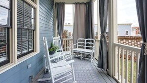 Eine überdachte Veranda mit weißen Schaukelstühlen, einem kleinen Tisch mit einem Blumentopf und Blick auf benachbarte Häuser.