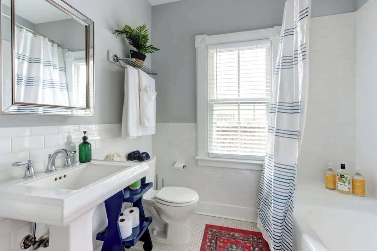 Une salle de bain avec un lavabo blanc, des toilettes, une fenêtre avec des stores, un rideau de douche et un miroir.