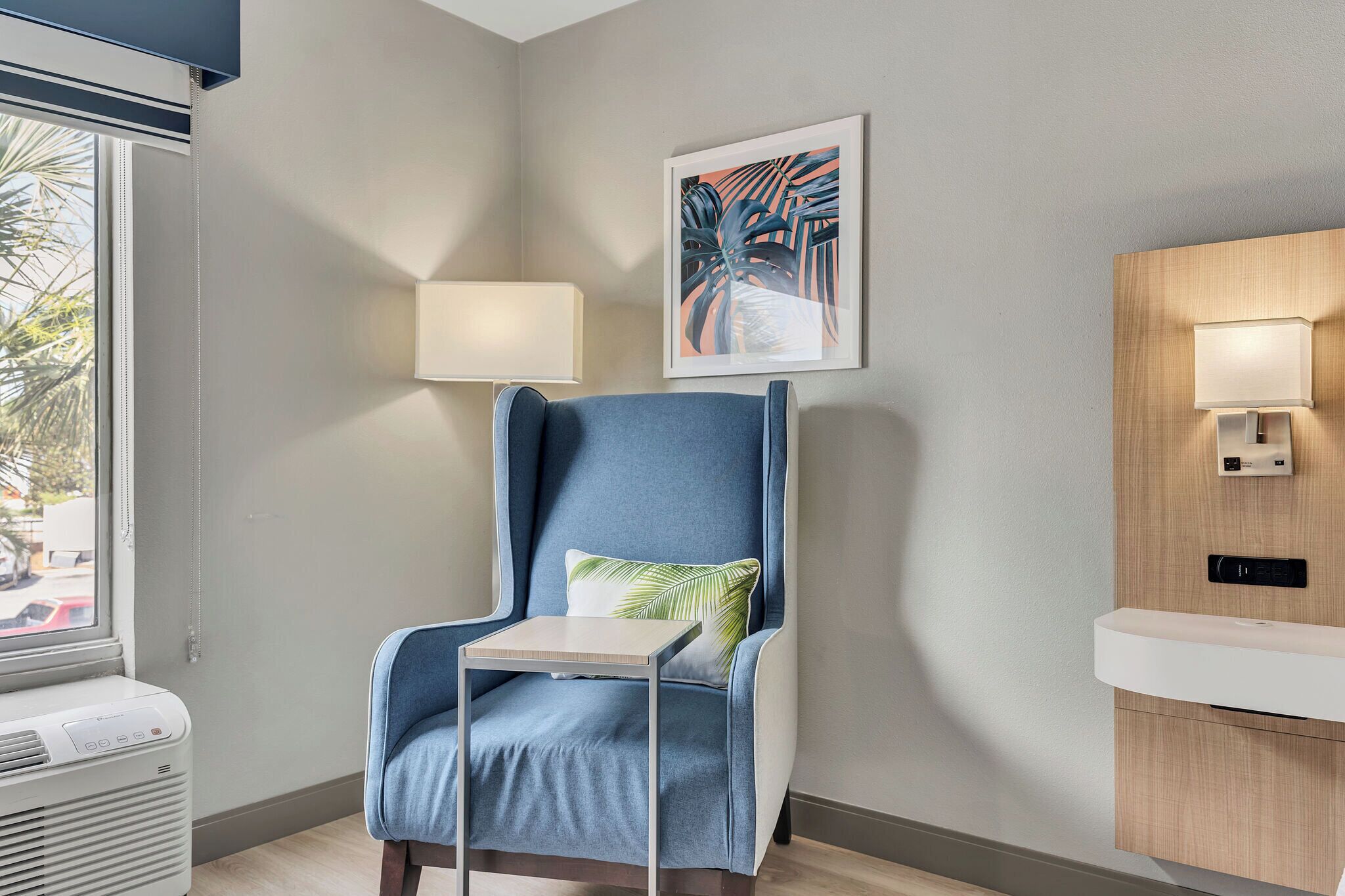 A modern room with a blue armchair, a small table, a wall-mounted lamp, a framed artwork, and a wooden cabinet with a built-in sink.