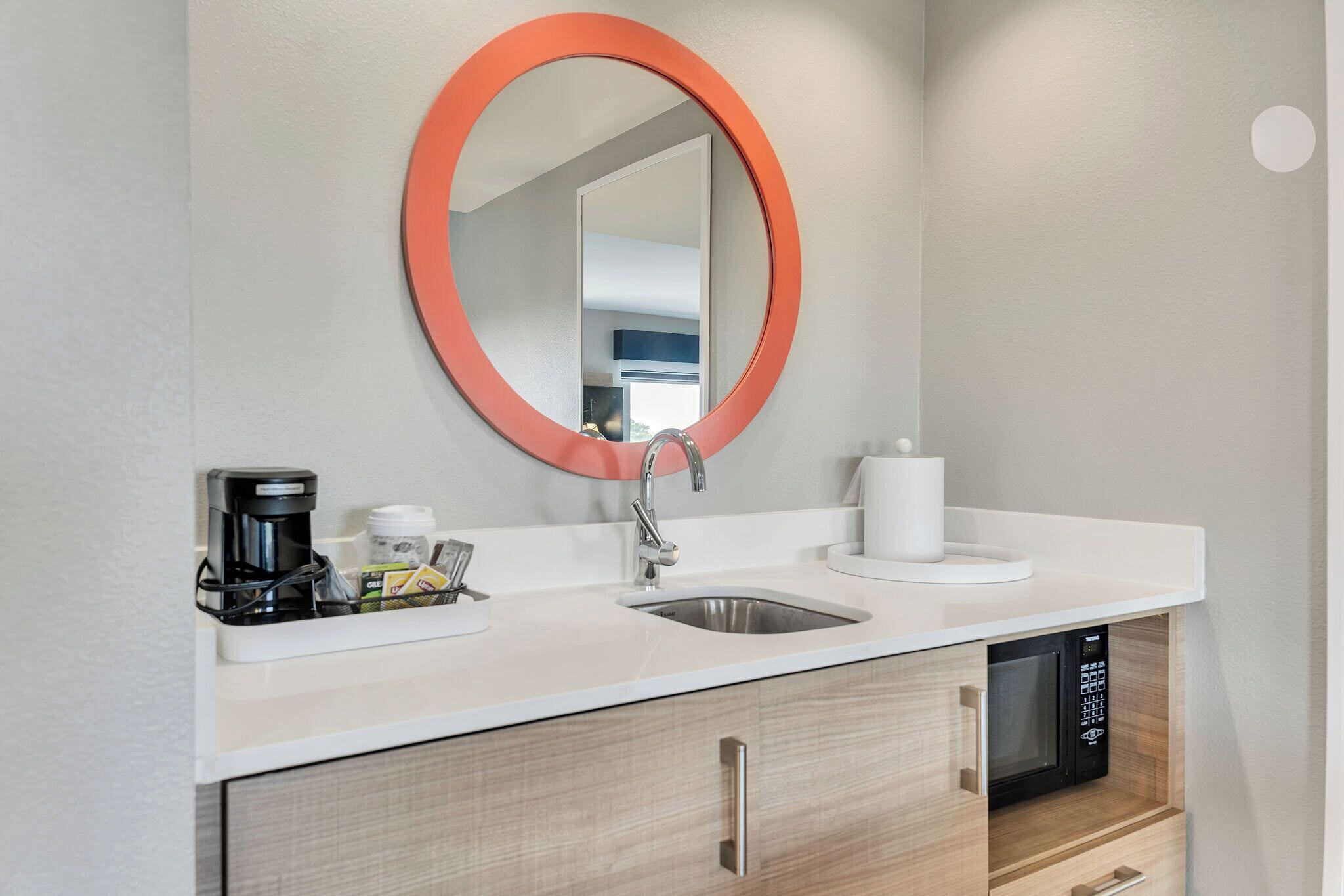 A modern bathroom with a sink, mirror, coffee maker, and a toilet paper holder.