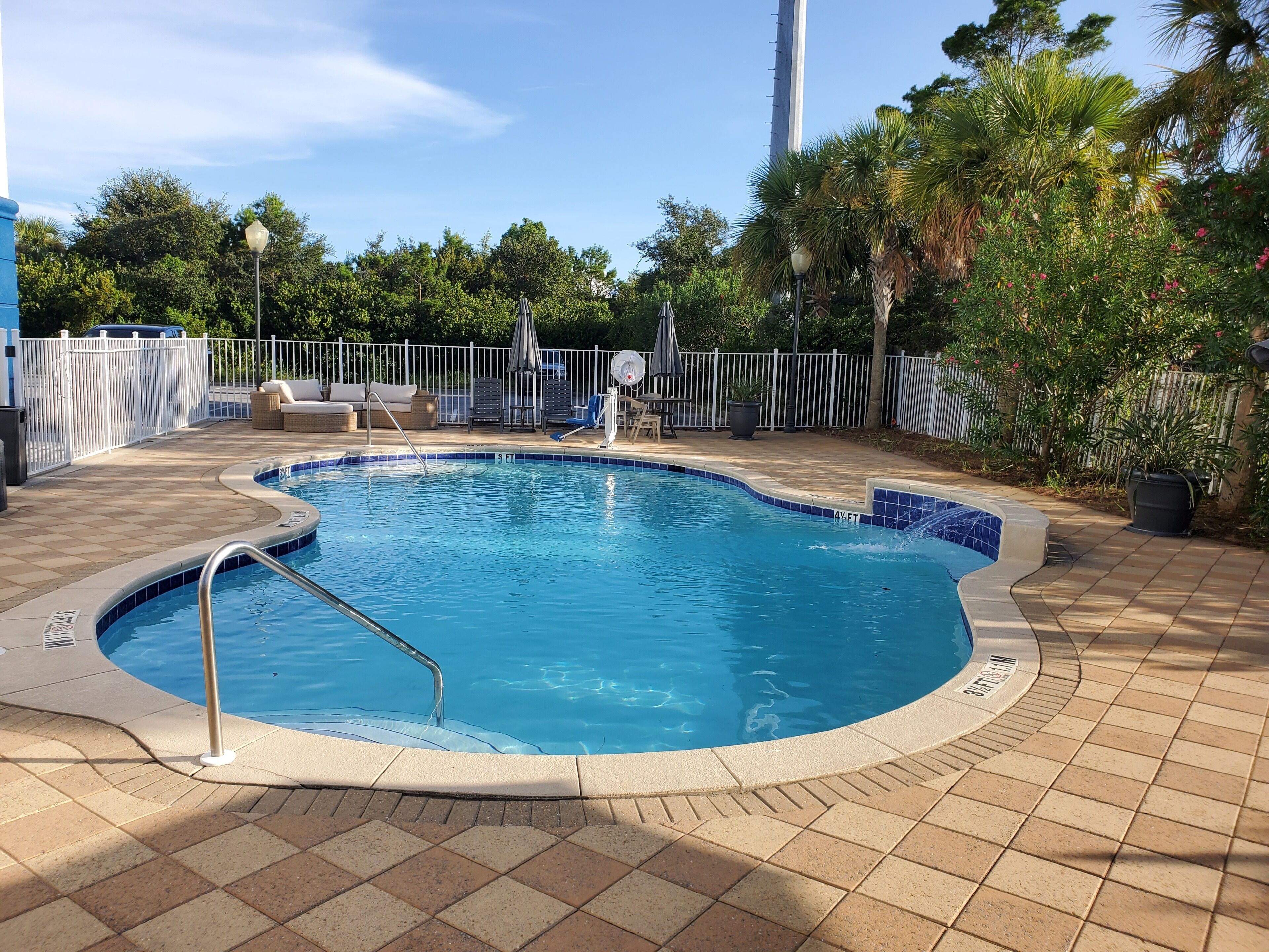 A swimming pool with a tiled edge, surrounded by a white fence, and a patio area with lounge chairs and umbrellas.
