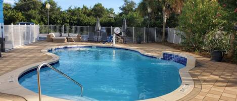 A swimming pool with a tiled edge, surrounded by a white fence, and a patio area with lounge chairs and umbrellas.