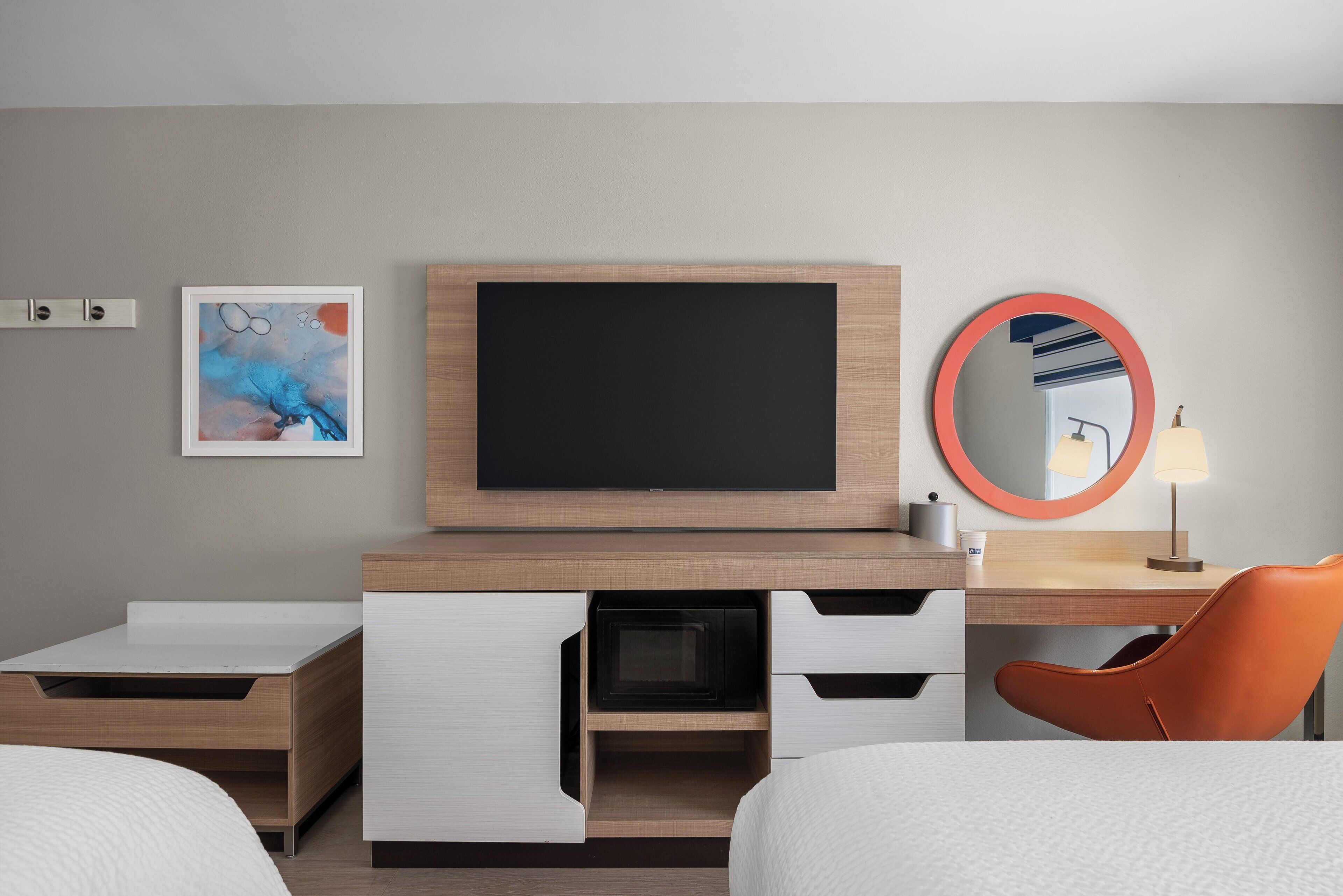 A modern hotel room with a flat-screen TV, a round mirror, a bed, and a desk with a lamp.