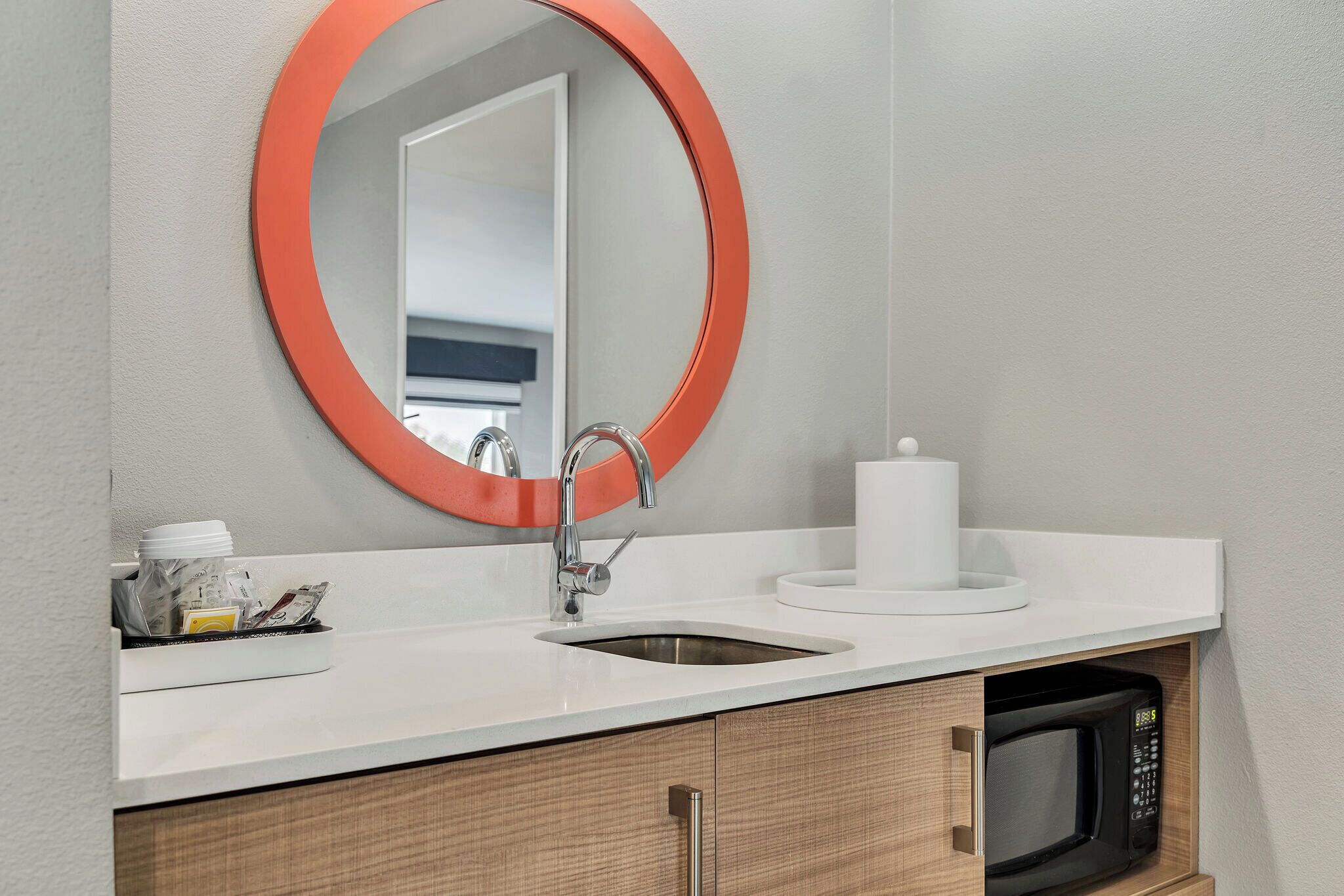 A bathroom with a white sink, a round mirror with an orange frame, a white candle, and a microwave built into the cabinetry.