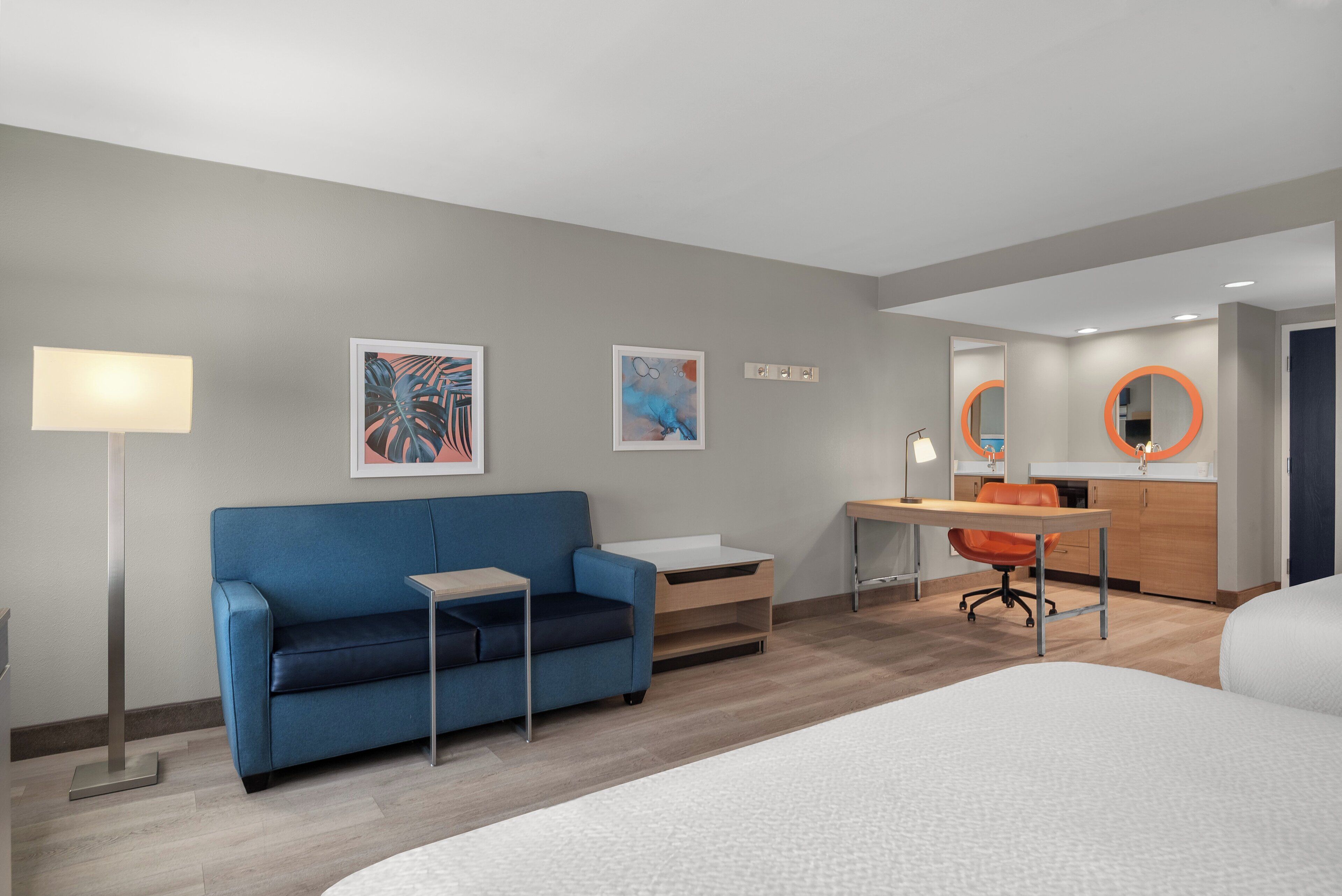 A modern hotel room with a blue sofa, a small desk, a bed, and a bathroom with a mirror and sink.