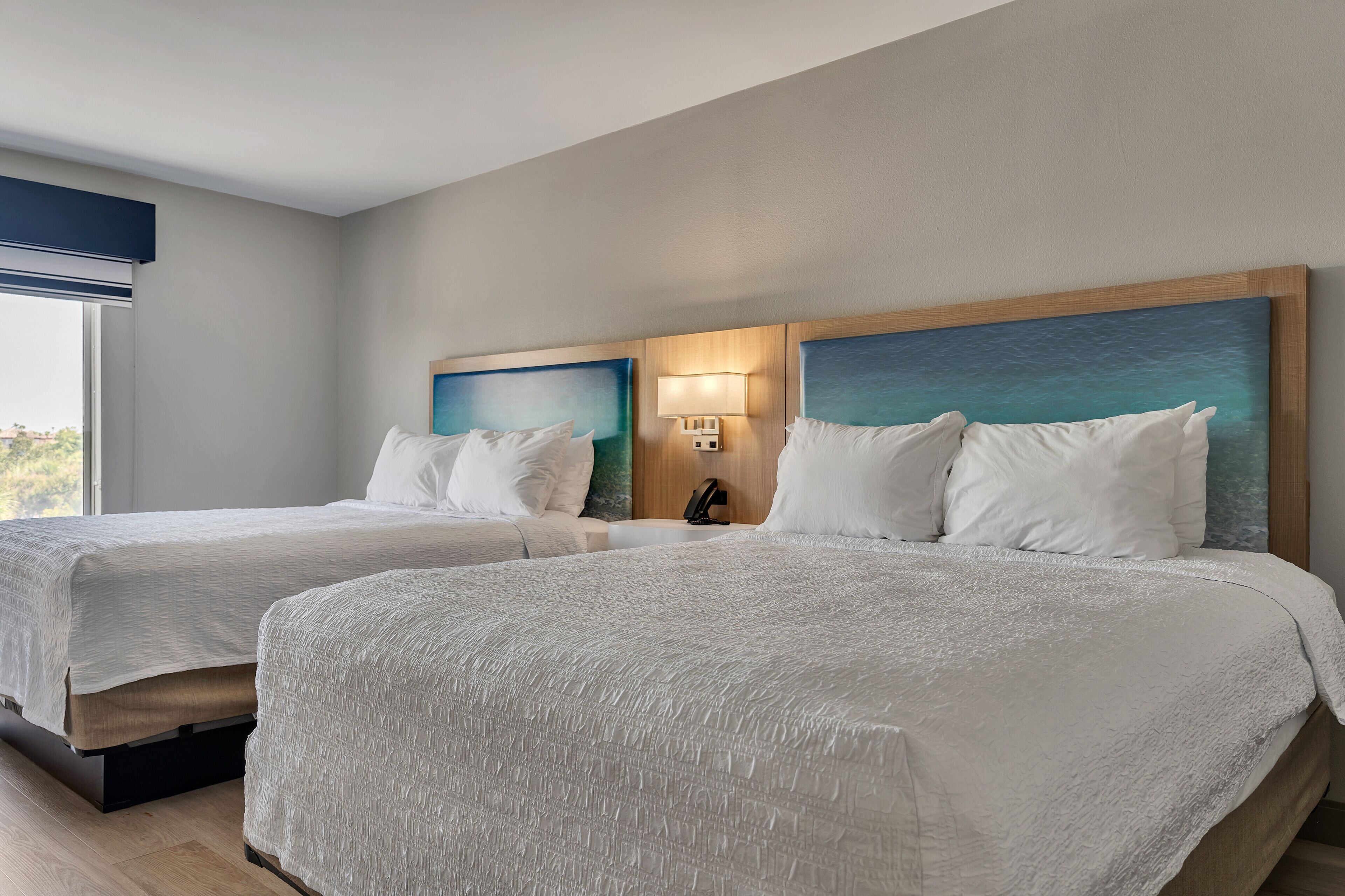 Habitación de hotel con dos camas, una mesita de noche, una lámpara y vistas al exterior.