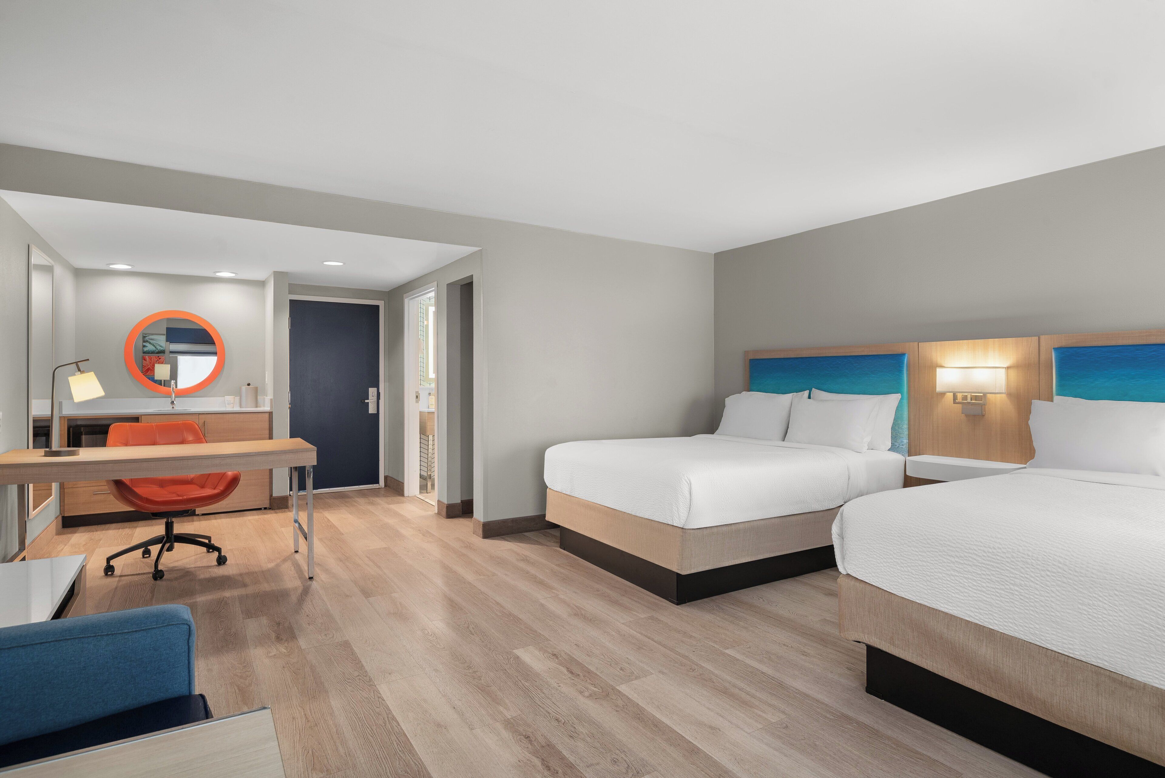 A hotel room with two beds, a desk with a chair, a mirror, and a lamp.