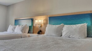 Habitación de hotel con dos camas, una mesita de noche con lámpara y una pared con vista al océano azul.