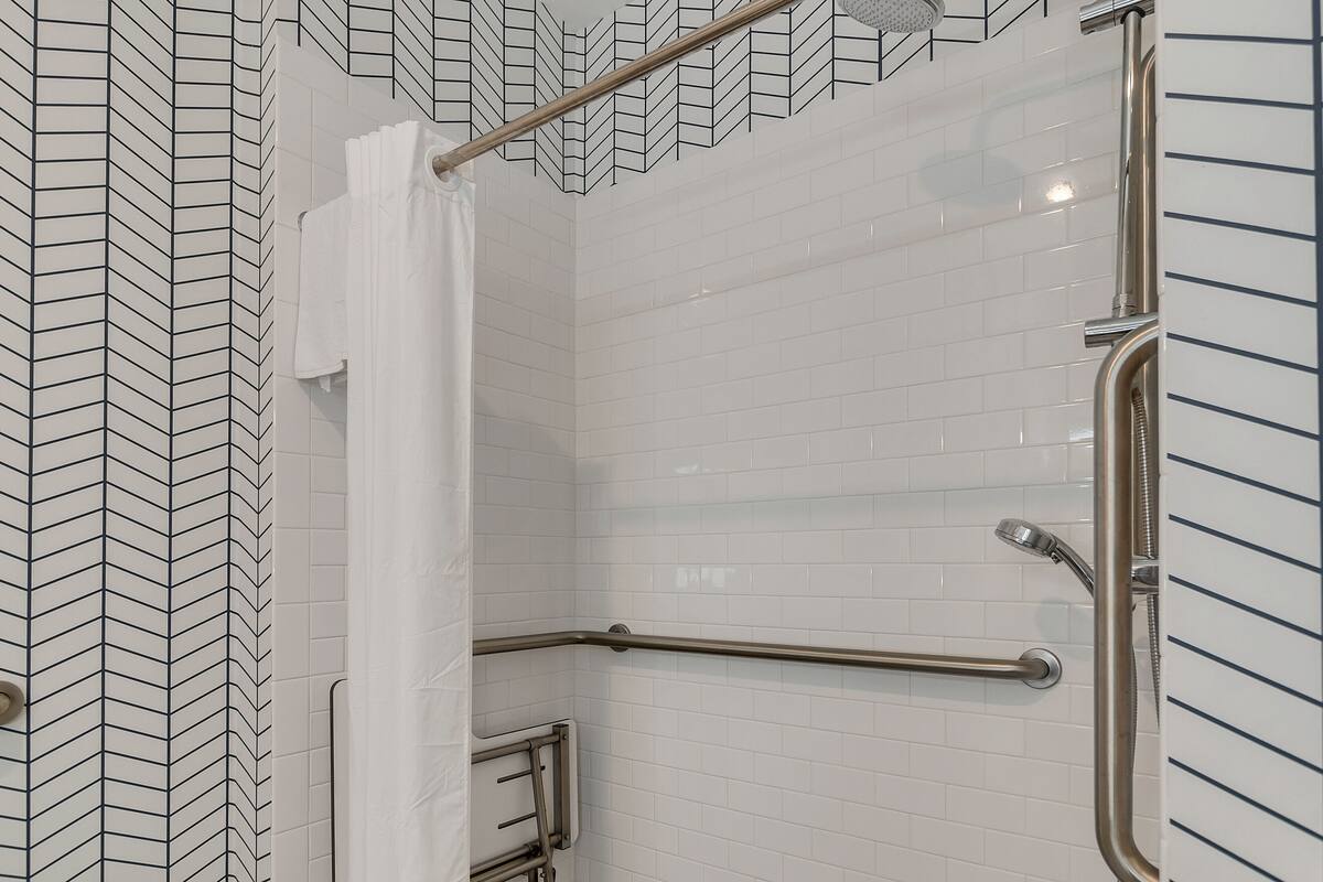 Zona de ducha con paredes de azulejos blancos, una barra de apoyo metálica y un cabezal de ducha.