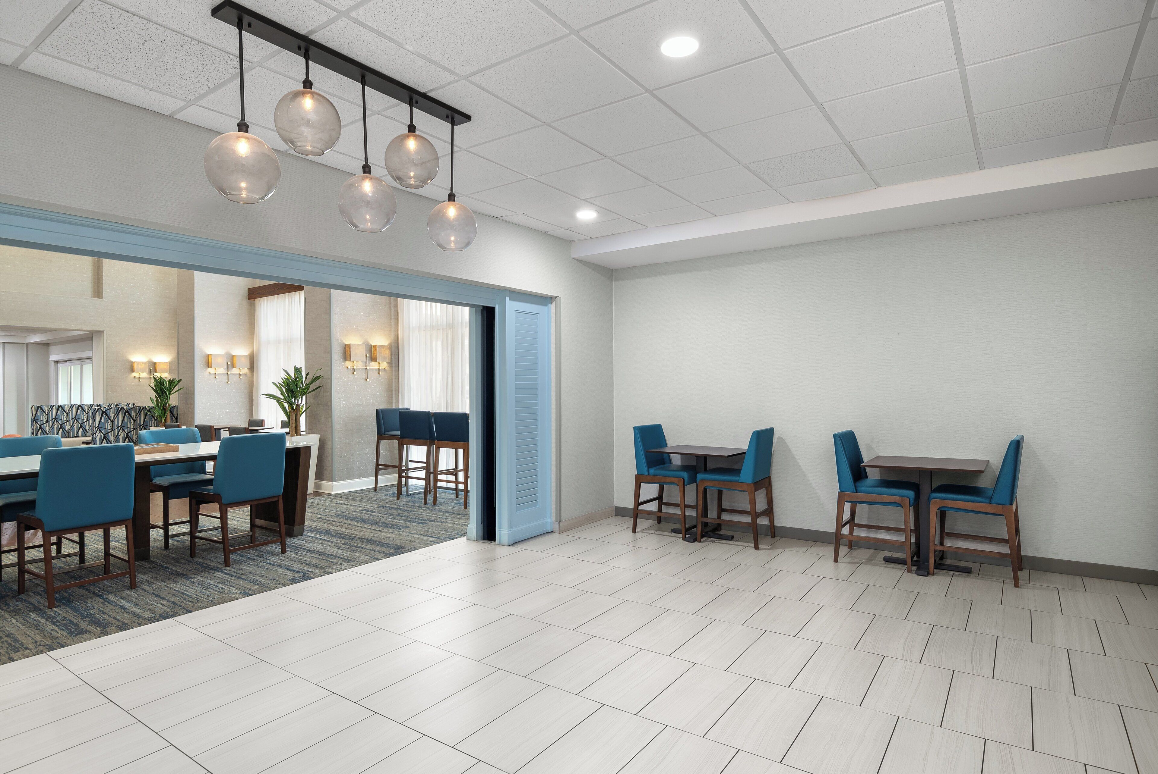 Un espace de restauration moderne dans un hôtel, caractérisé par un grand espace ouvert, une salle à manger séparée avec des chaises bleues et un plafond orné de luminaires sphériques suspendus.