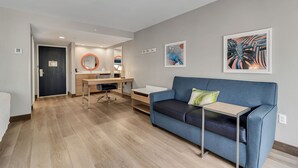 Una habitación de hotel moderna con un sofá azul, una mesita, un escritorio con una silla y obras de arte en las paredes.