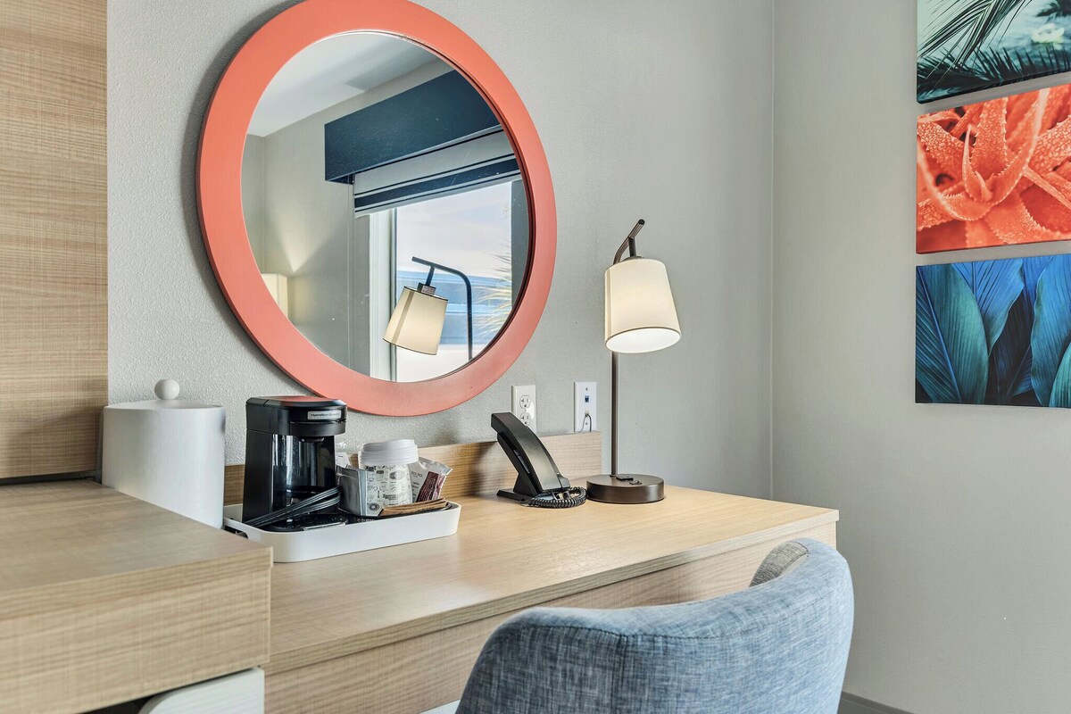 A hotel room with a wooden desk, a coffee maker, a telephone, a lamp, and a mirror with a red frame.
