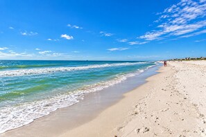Une plage à l’eau d’un bleu limpide, avec un rivage sablonneux et quelques personnes qui se promènent.