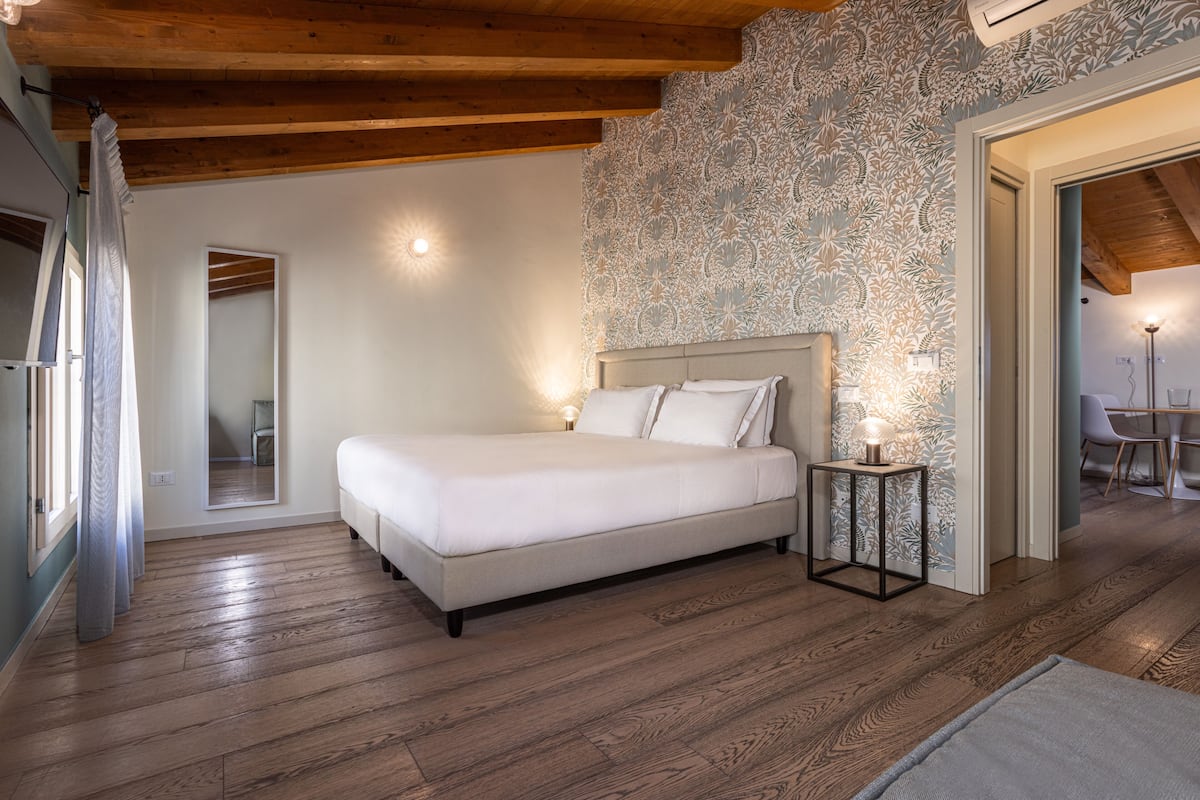 A bedroom with a large bed, wooden flooring, a patterned wall, and a small bedside table with a lamp.