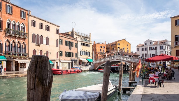 A canal with boats, a wooden post, and buildings with balconies and shops. - Palazzina Riello (Venice)