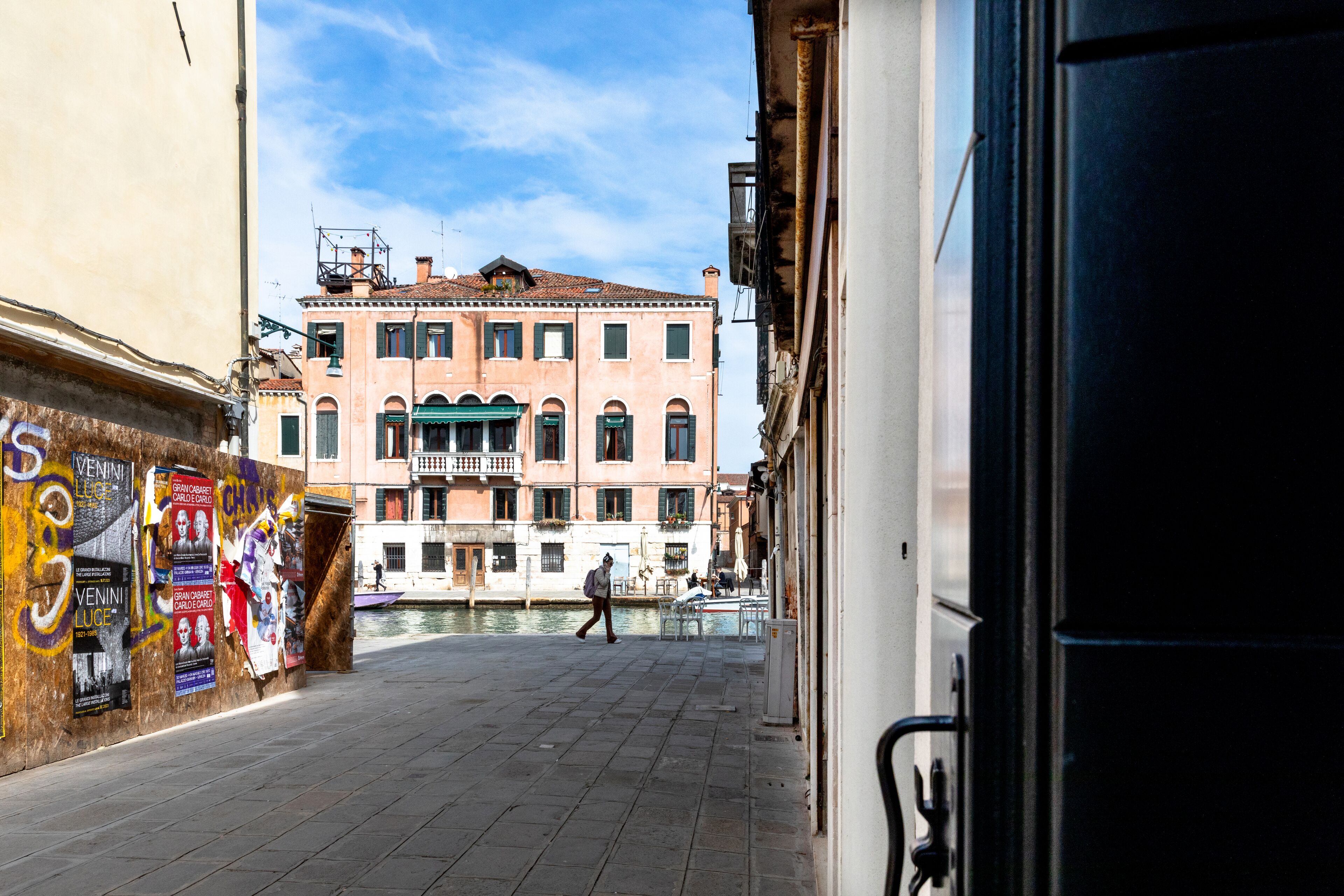 A narrow street with a canal, buildings with balconies, and a person walking.
