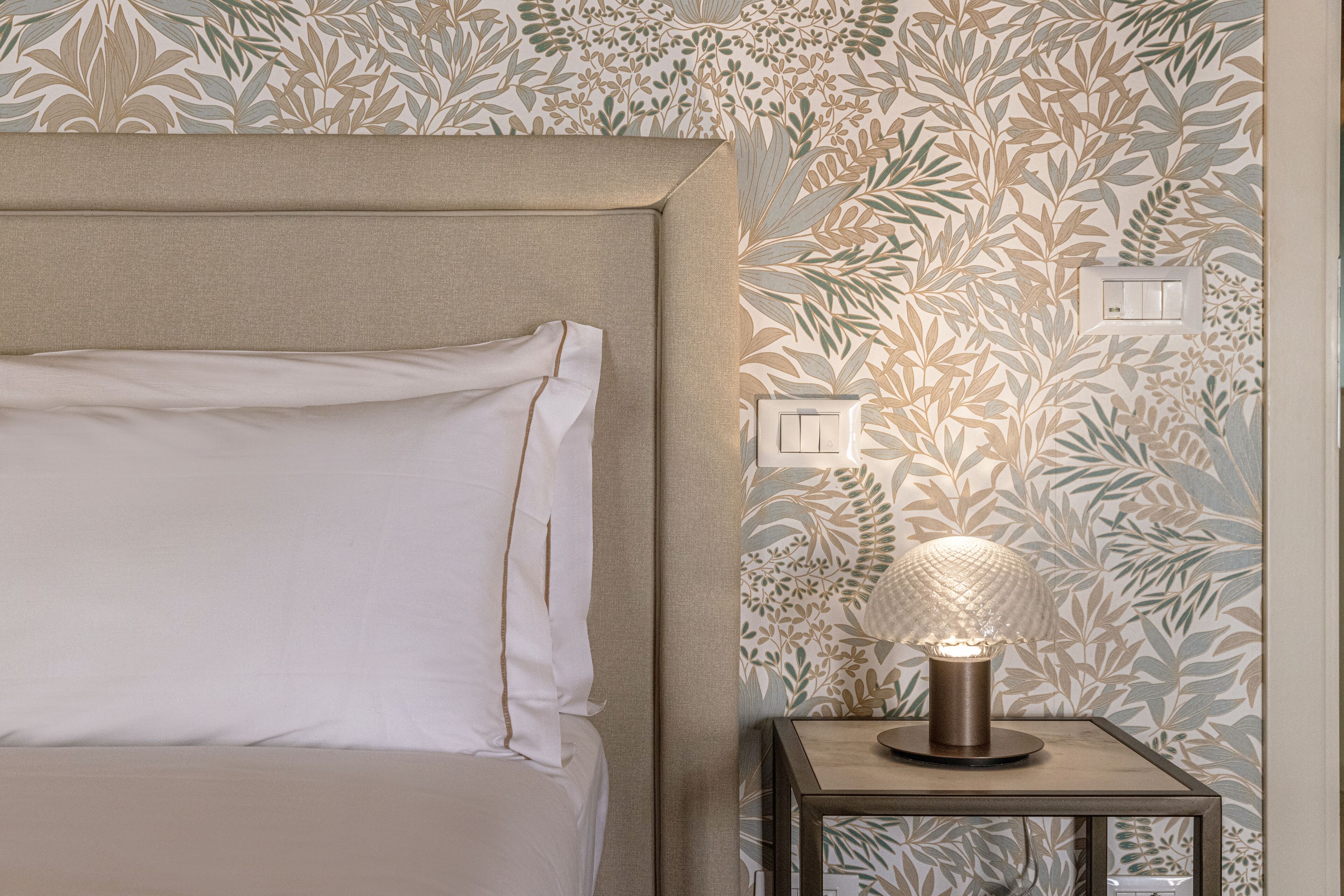 Un lit bien fait, avec une tête de lit, une lampe de chevet et un mur orné d’un motif végétal.