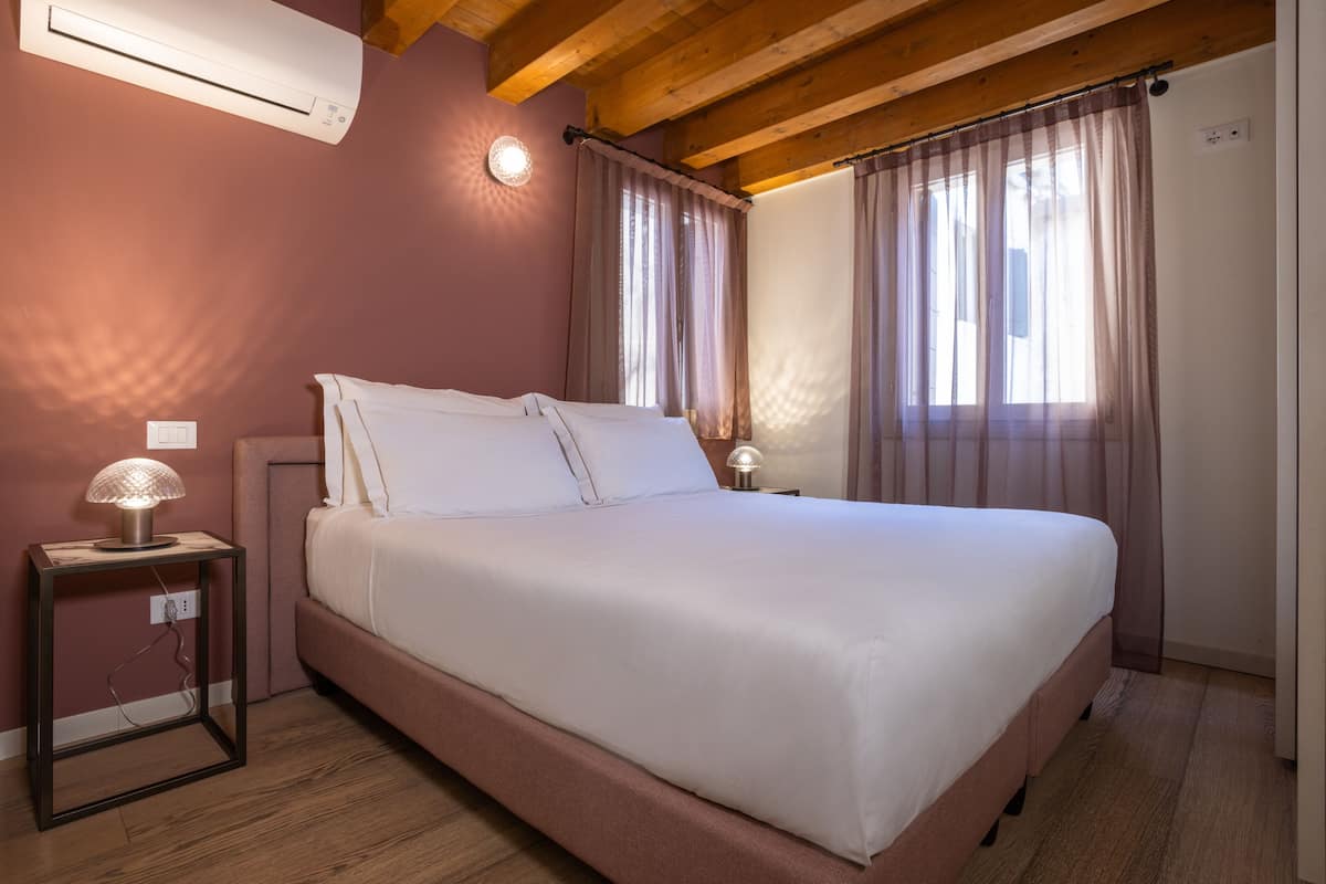 A bedroom with a large bed, two bedside tables with lamps, a wall-mounted air conditioner, and a window with curtains.