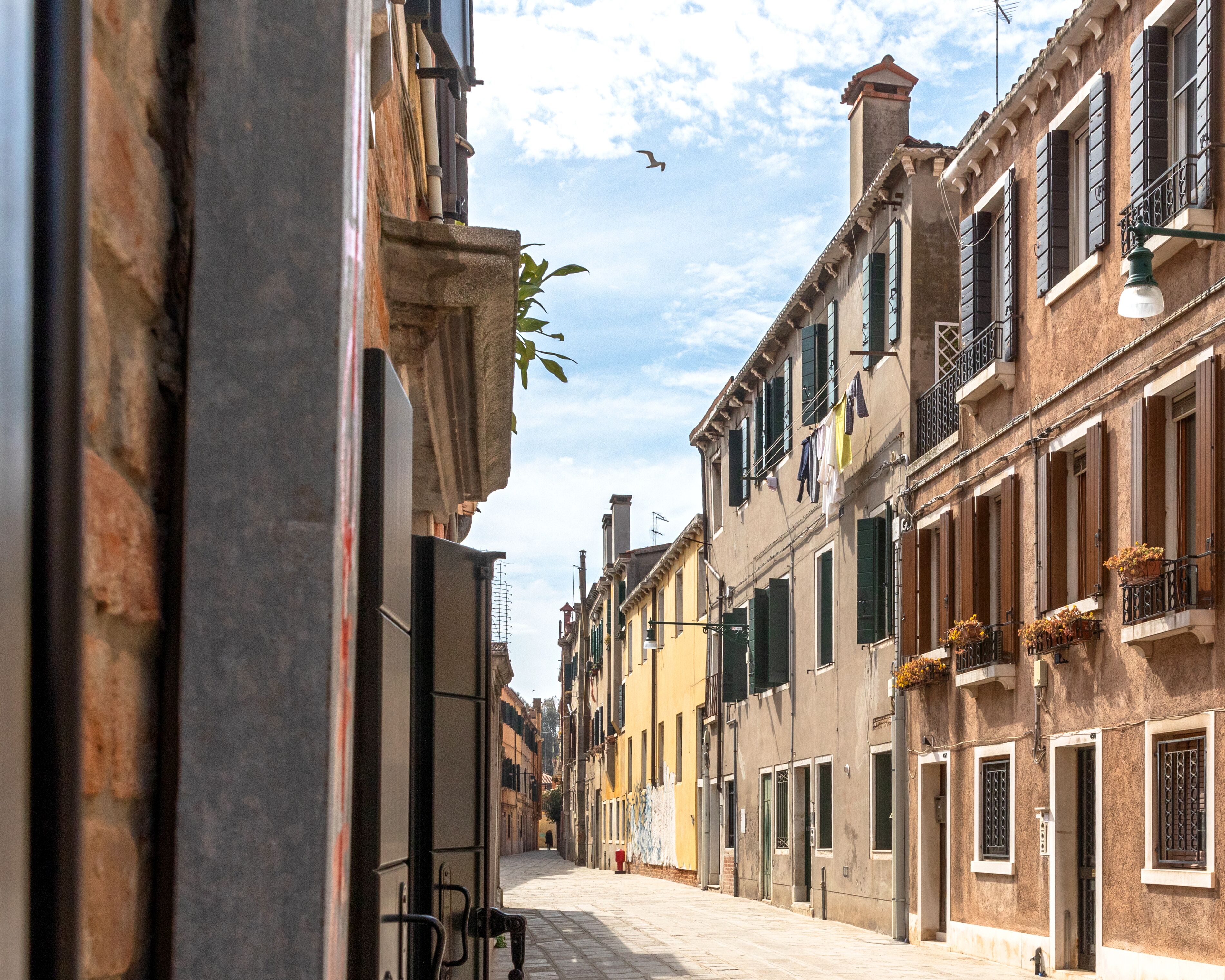 A narrow street lined with historic buildings, featuring balconies with flowers, a clear sky, and a bird in flight.