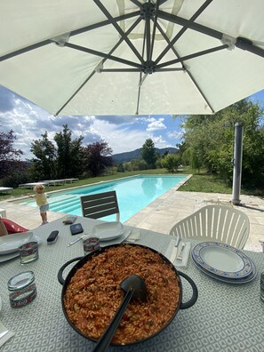 Un espace de restauration en plein air avec vue sur la piscine, une table sur laquelle se trouve un plat, et un parasol blanc.