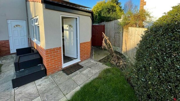 Ein kleines, eingeschossiges Haus mit weißer Tür, einer kleinen Veranda und einem Sockel aus Ziegeln.