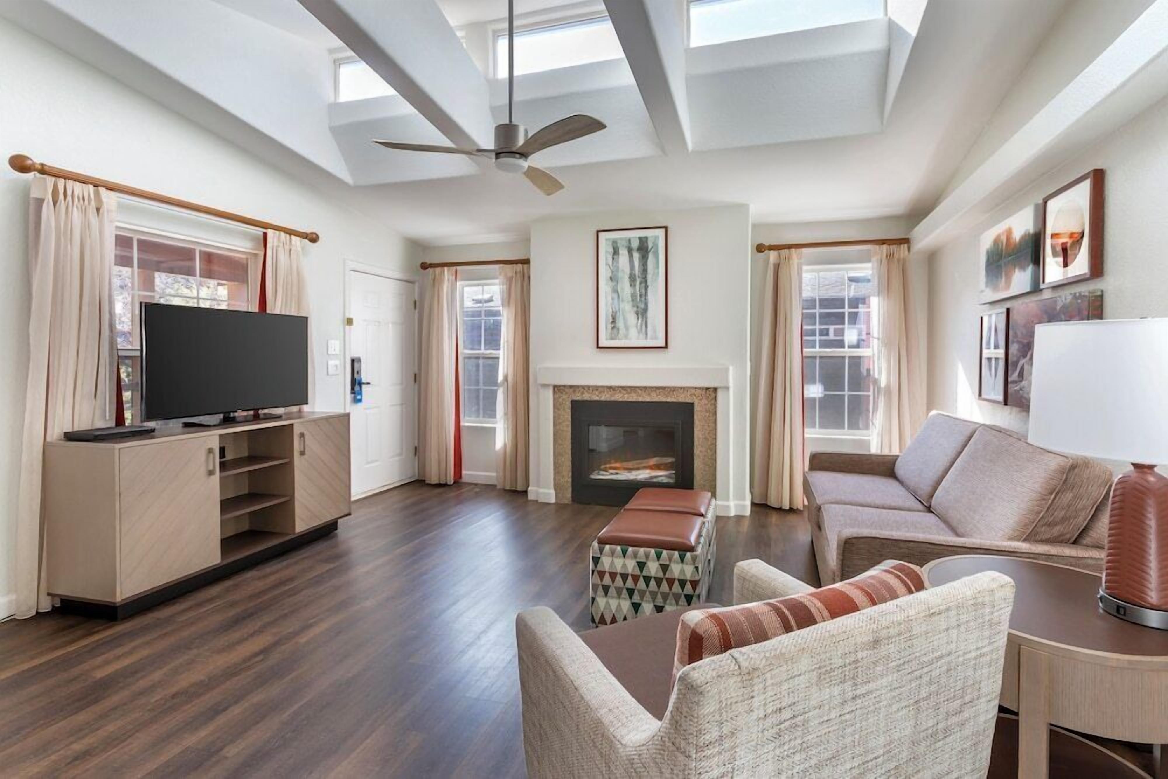 A spacious living room with a flat-screen TV, a fireplace, a ceiling fan, and a large window providing natural light.