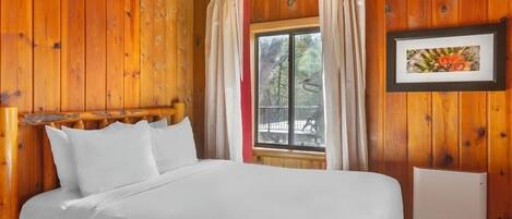 A wooden cabin bedroom with a bed, a window with curtains, and a framed picture on the wall.
