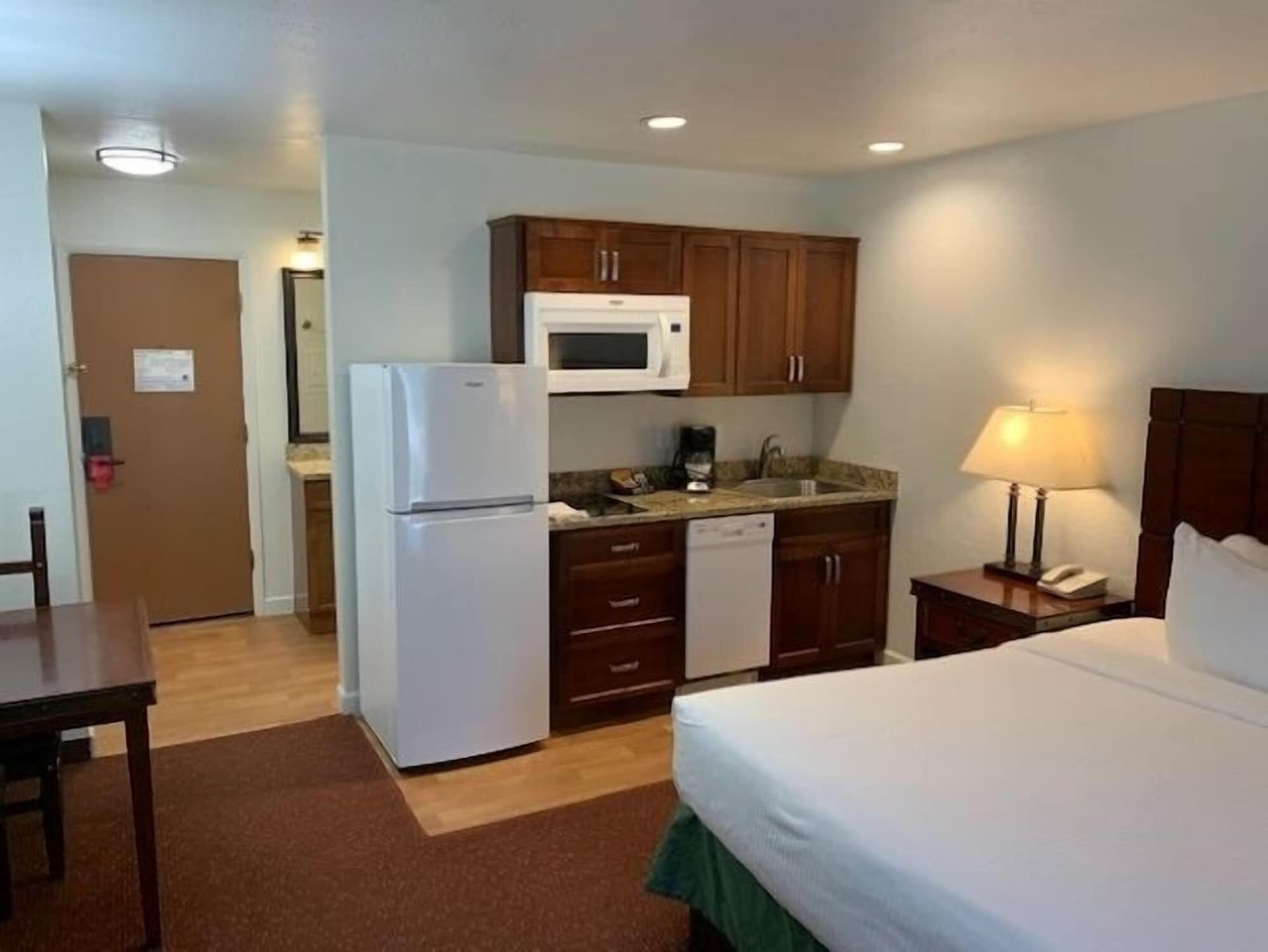 Une chambre d’hôtel comprenant un lit, une petite kitchenette équipée d’un réfrigérateur, d’un four à micro-ondes et d’un lave-vaisselle, une table de chevet avec une lampe, et une porte donnant accès à la salle de bain.