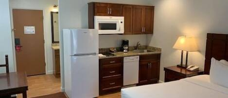 Une chambre d’hôtel comprenant un lit, une petite kitchenette équipée d’un réfrigérateur, d’un four à micro-ondes et d’un lave-vaisselle, une table de chevet avec une lampe, et une porte donnant accès à la salle de bain.