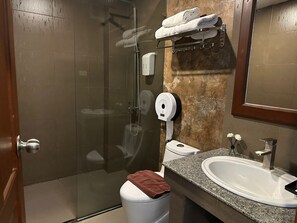 Une salle de bain moderne avec une douche à parois de verre, des toilettes blanches, un lavabo surmonté d’un plan de travail en granit et un porte-serviettes avec des serviettes pliées.