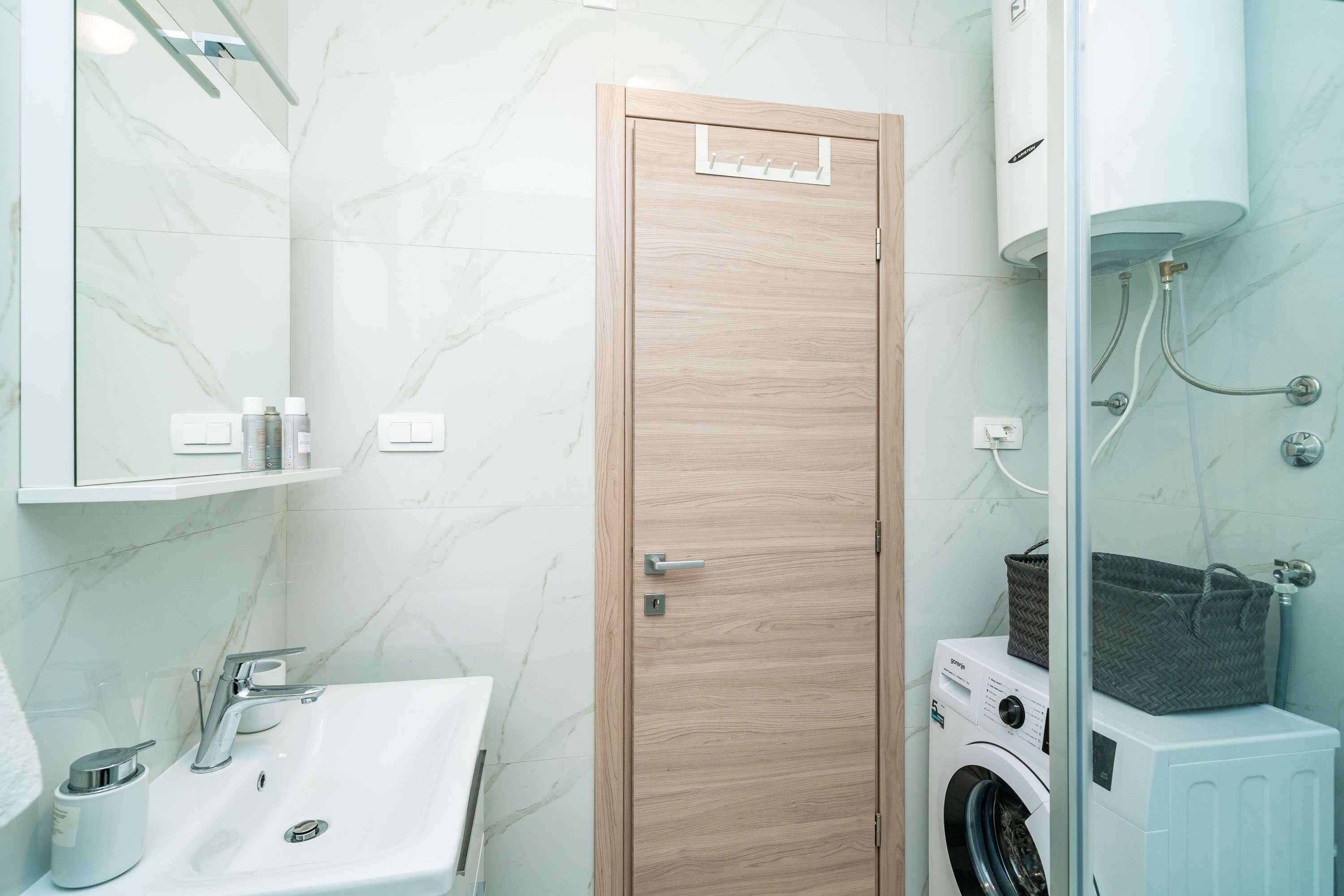 Une salle de bain moderne avec un mur en marbre, une porte en bois, un lavabo avec un robinet, un lave-linge et une cabine de douche en verre.